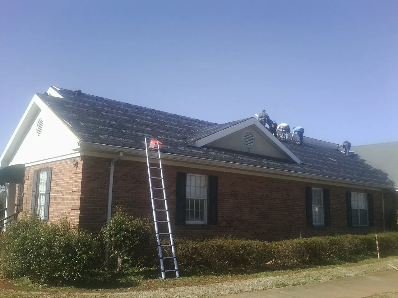 Crew tearing shingles off a brick ranch roof with a ladder set — Metal Roof Repair work by our crew in Colora