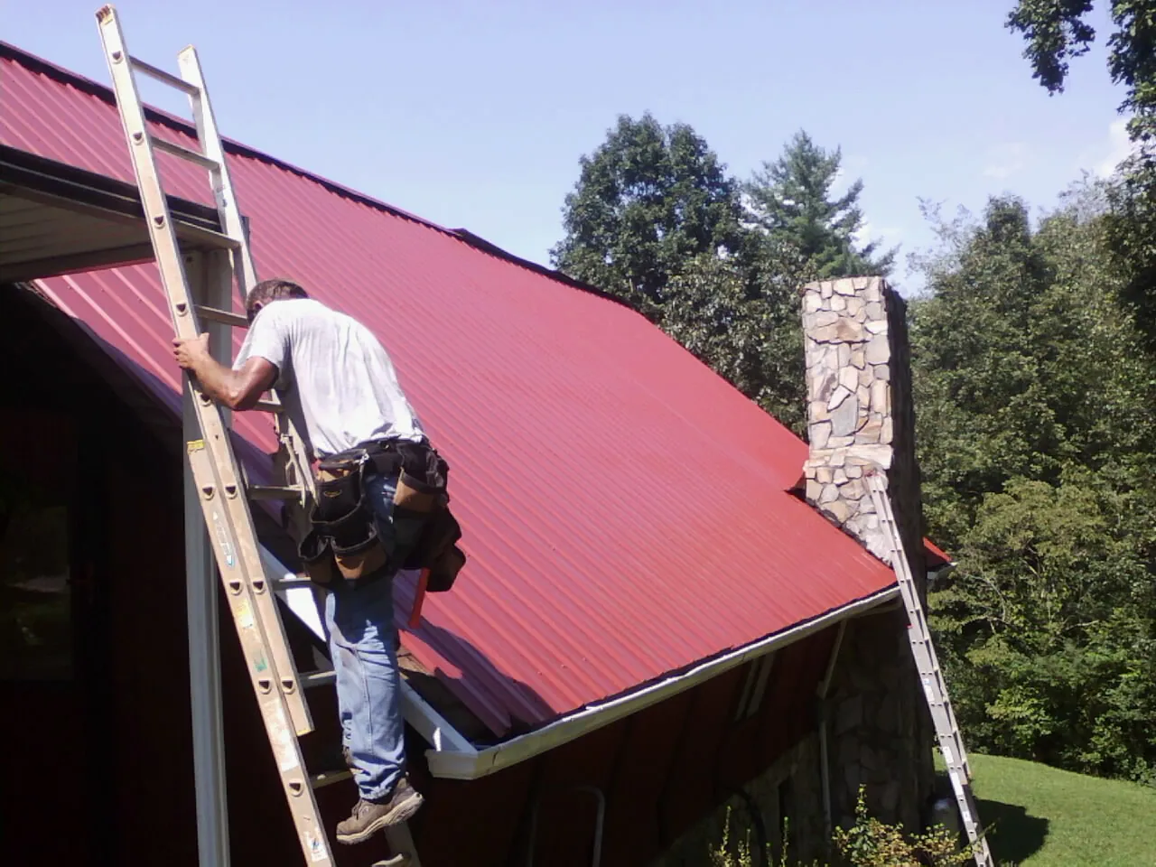 Roofer on a ladder beside a red metal roof and stone chimney — Soffit & Fascia Repair by Roofing Brothers in Colora